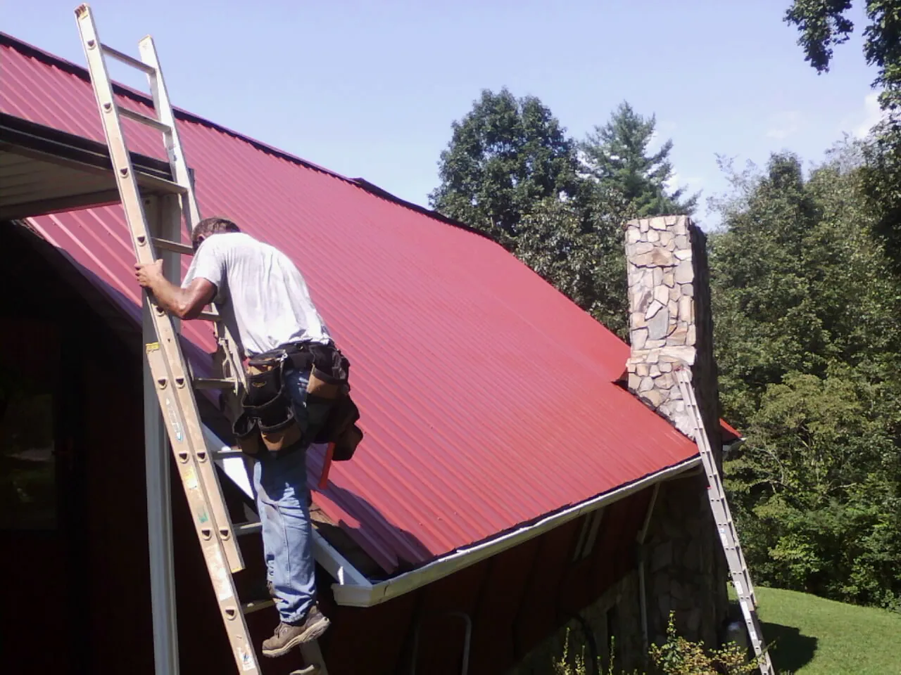 Roofer on a ladder beside a red metal roof and stone chimney — Soffit & Fascia Repair by Roofing Brothers in New Philadelphia